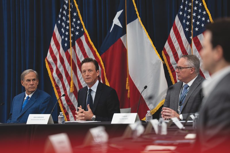 Governor Greg Abbott, White House National Cyber Director Sean Cairncross, and Texas Cyber Command Chief TJ White seated at a table before U.S. and Texas flags at the Project Watershed 250 launch in San Antonio.